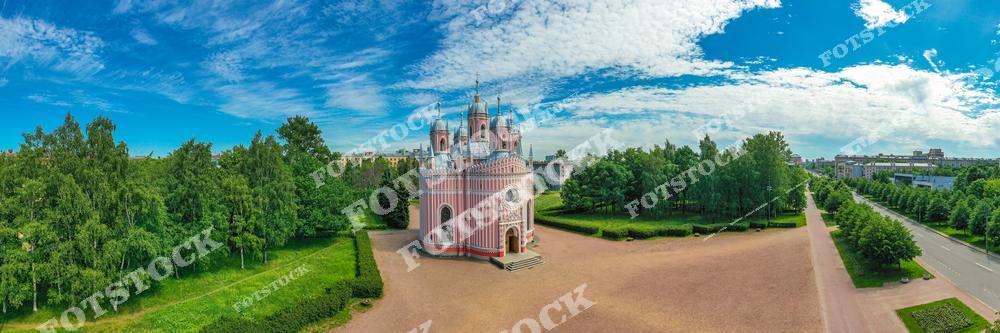 На фотографии изображена Чесменская церковь — один из самых необычных храмов Санкт-Петербурга, выполненный в стиле неоготики с элементами барокко. Храм находится в Московском районе и окружён парковой зоной. Его розово-белый фасад и изящные формы выделяются на фоне зелени и голубого неба. Просторная площадь перед церковью подчёркивает её симметрию и красоту. Снимок сделан с воздуха в солнечный летний день.